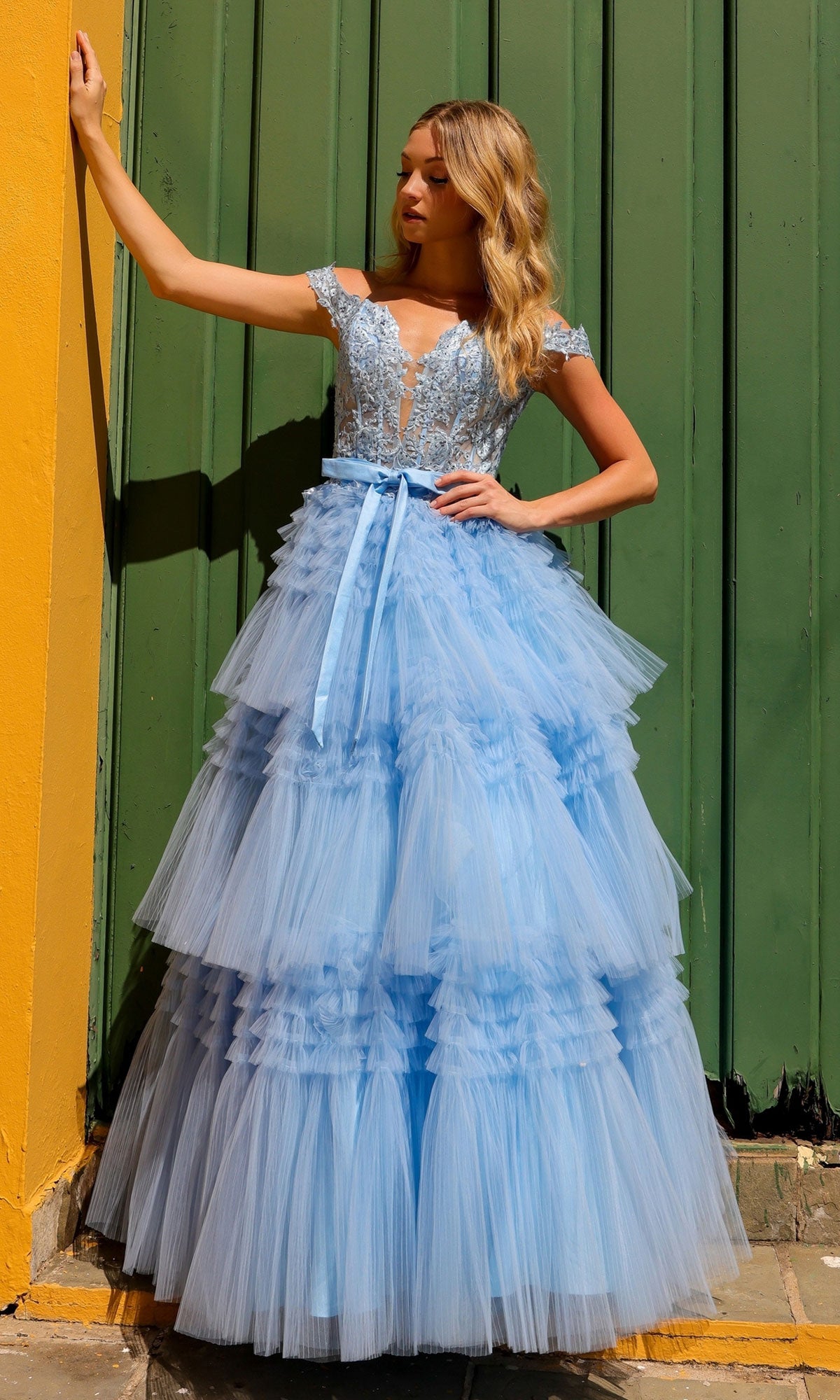 A woman stands by a yellow wall and green doors wearing the Nox Anabel Designer Ruffled Prom Ball Gown E1293—a light blue, off-the-shoulder tiered tulle dress with beaded lace embroidery and a satin ribbon belt.